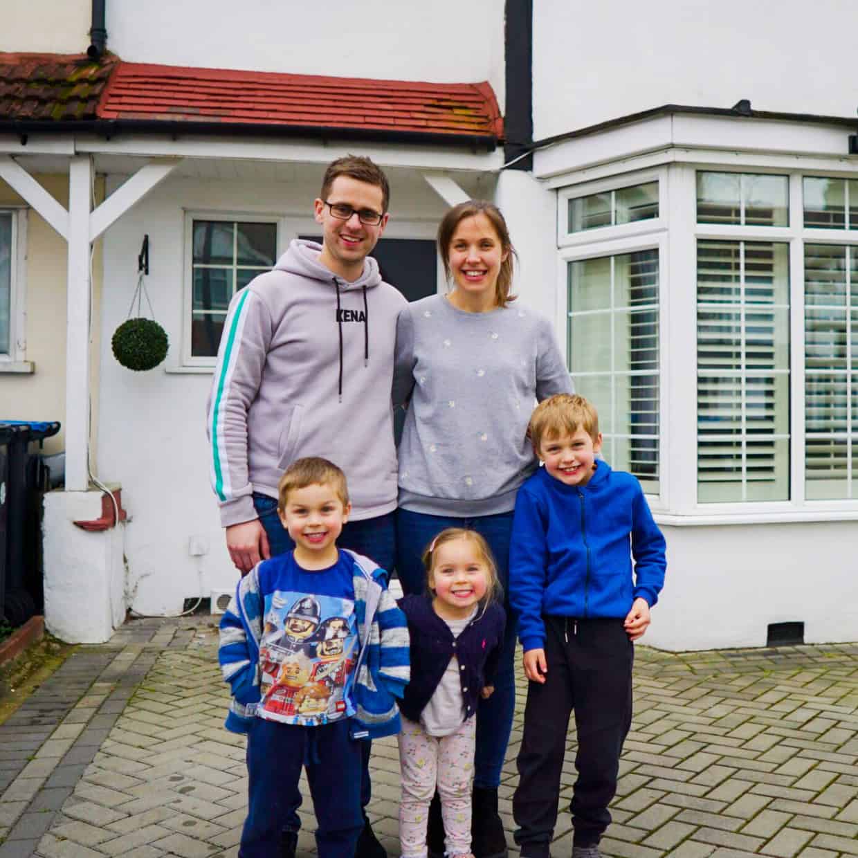 Smiling family of five standing outside their home, with parents behind and three young children in front on a paved driveway.