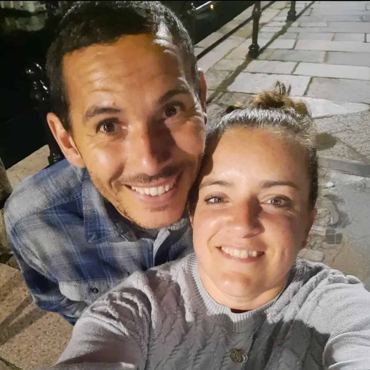 Smiling couple taking a selfie at night on a paved walkway beside water with metal railings in the background.