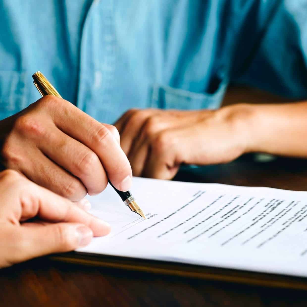 Close-up of a person signing a document with a fountain pen, while another person points at the paper, suggesting guidance or explanation.