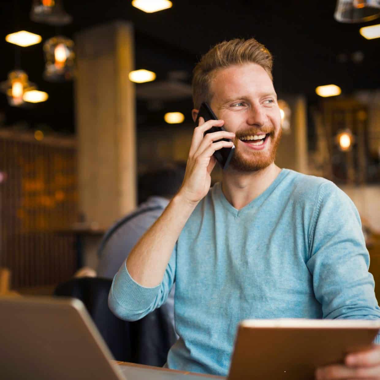 "Smiling man on phone call, holding tablet and working in a café.