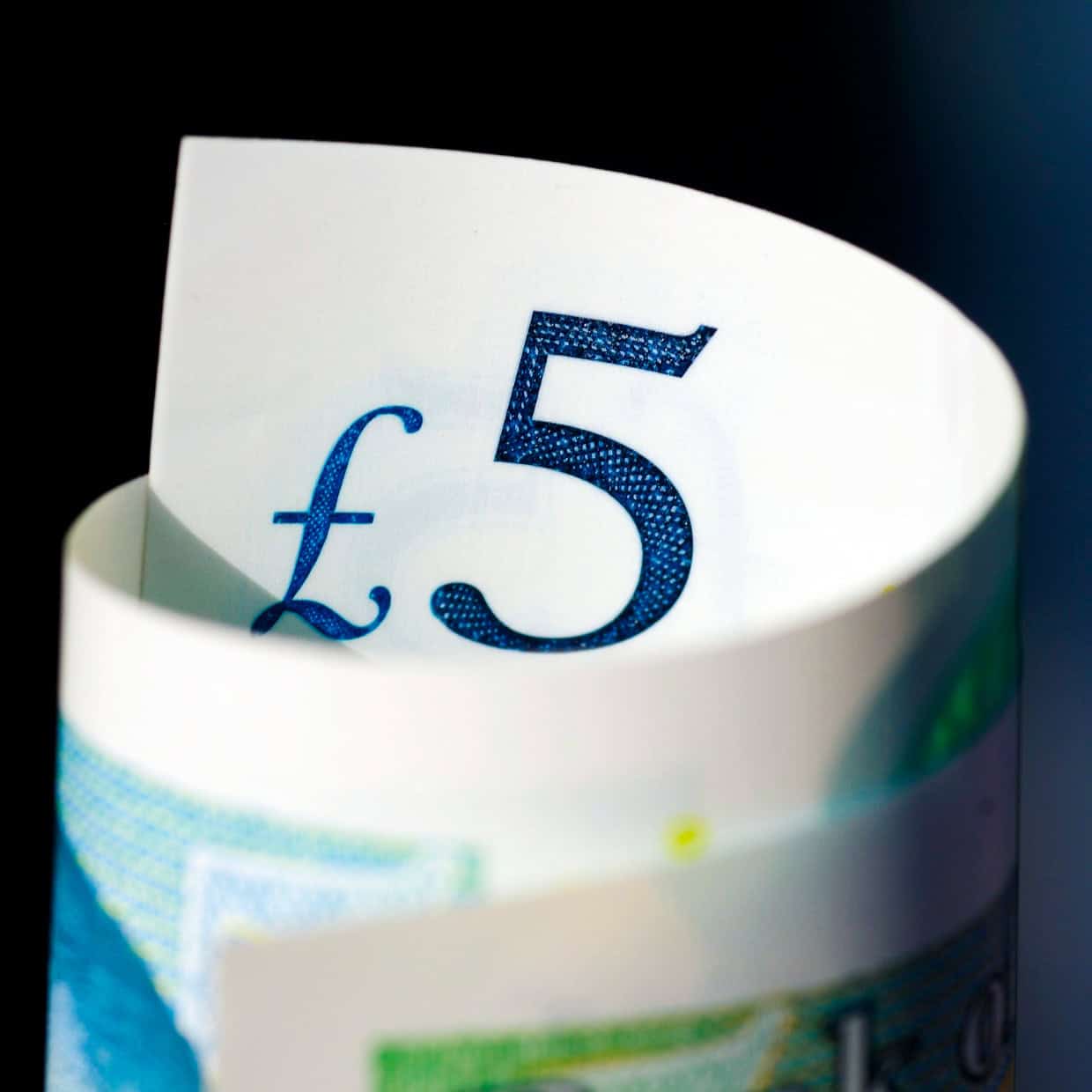 Close-up of a UK five-pound note showing the blue £5 symbol against a dark background.