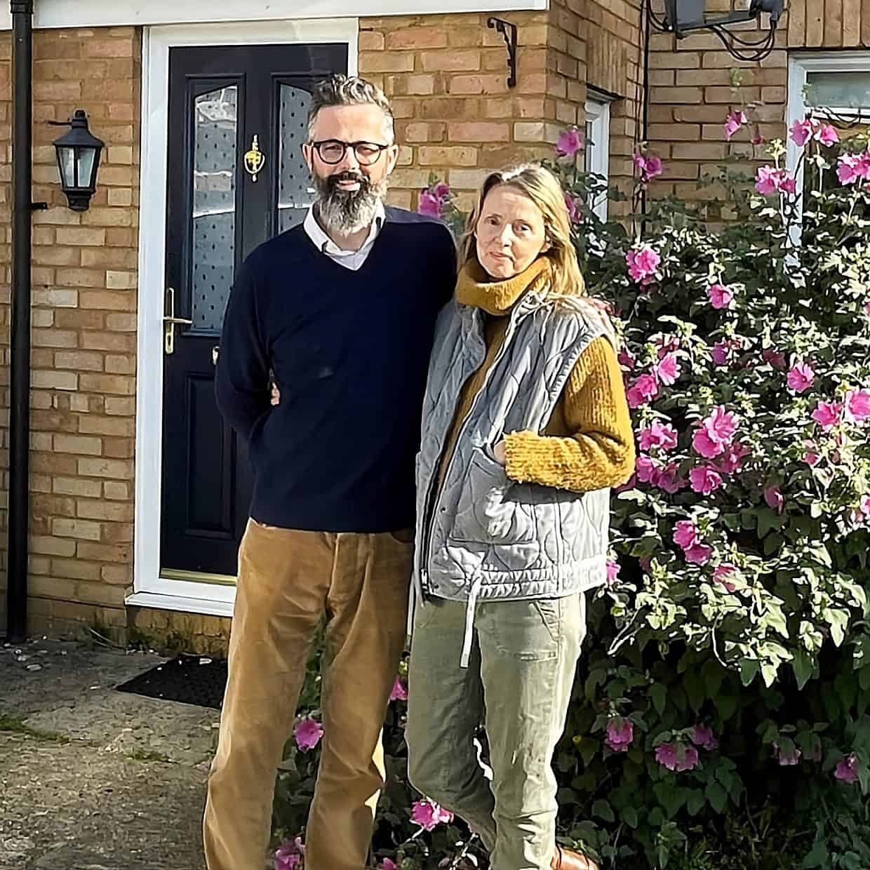 Couple standing together outside a brick house with a black front door, next to a car and a large bush covered in pink flowers