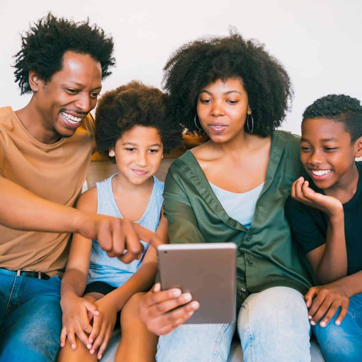 Happy family of four sitting on a sofa, looking at a tablet together and smiling.